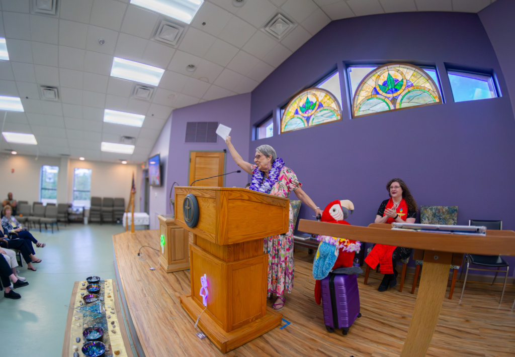 Pat Felton holding the pledge-drive announcement with Jerry the parrot mascot; Rev. Kristina seated behind in the sanctuary