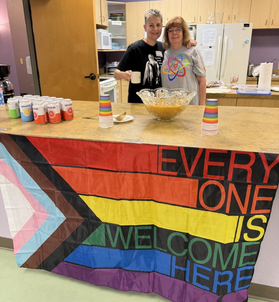 Two members behind the refreshment counter, draped with a Progress Pride flag that reads "Every One Is Welcome Here."