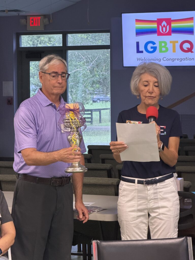 Two members at the front of the room beneath an LGBTQ Welcoming Congregation slide; one holds a glass trophy while the other reads from a page.