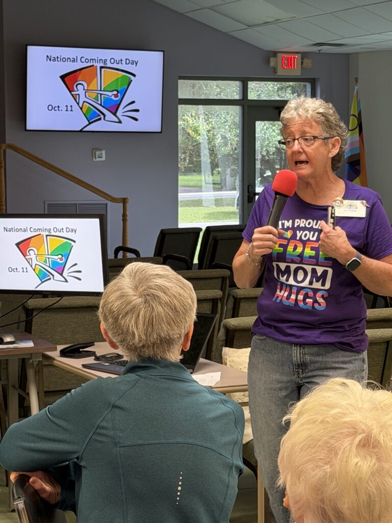 A member in a purple T-shirt speaks at the microphone beside a National Coming Out Day slide.