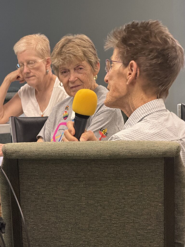 Three members talking at a table; one holds a microphone.