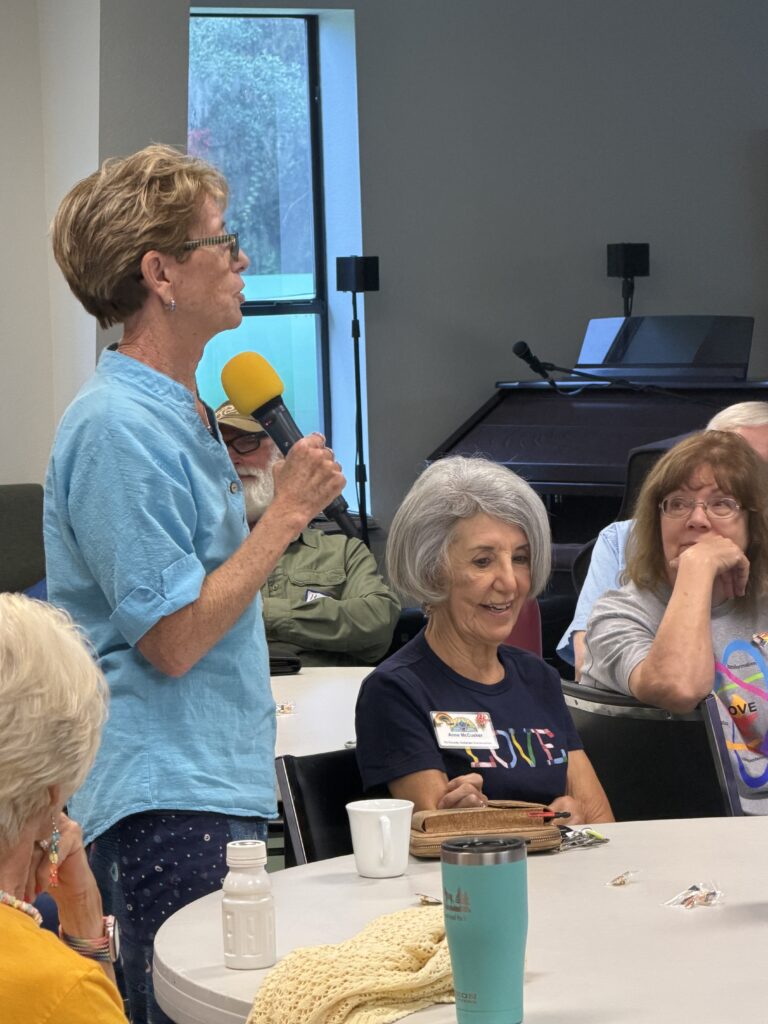 A member in a light blue shirt speaks into a microphone while others listen at their tables.