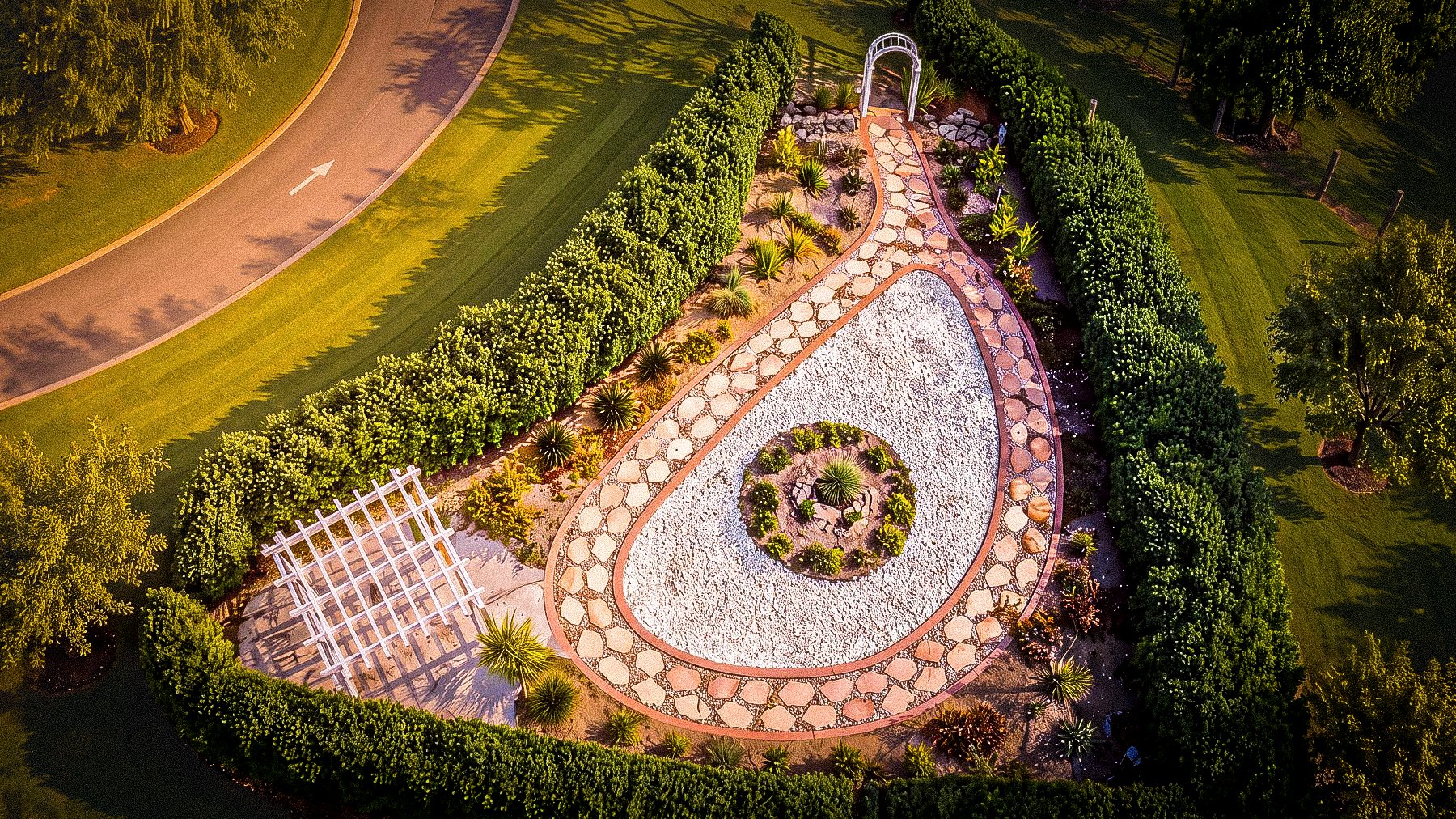 Aerial view of the TriUU Memorial Garden: a teardrop-shaped flagstone path around a bed of white shell and a planted circle, enclosed by clipped hedges, with a white pergola and arch.