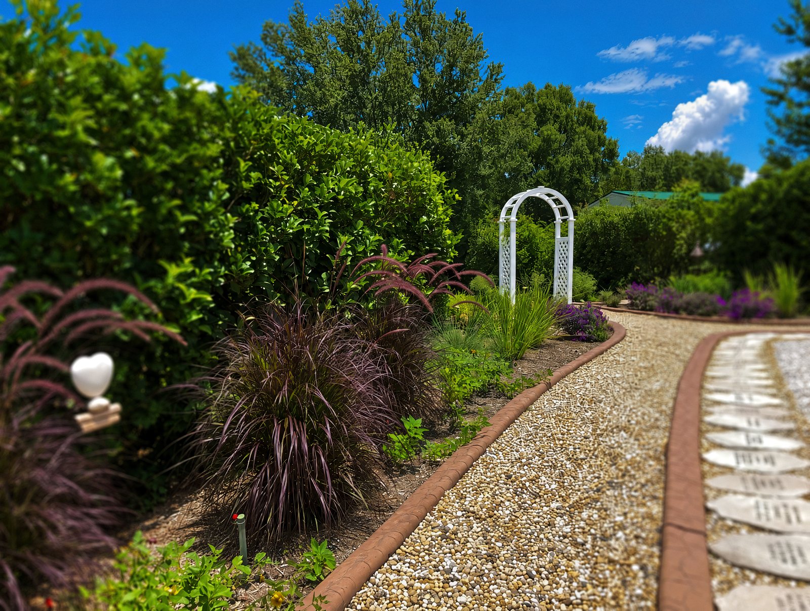 A gravel path in the TriUU Memorial Garden, edged with a row of engraved memorial stones, leading to a white arch between hedges and ornamental grasses.