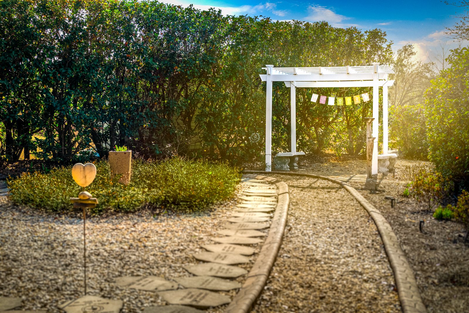 A gravel path lined with memorial stones curves through the TriUU Memorial Garden toward a white pergola hung with prayer flags, in late-afternoon sun.