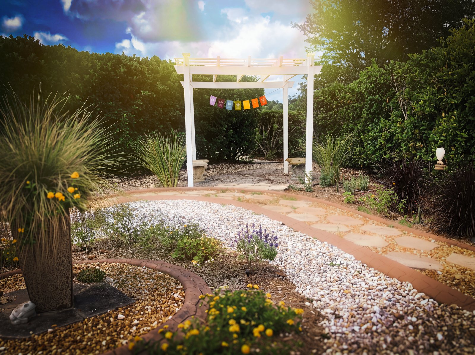 The TriUU Memorial Garden in warm afternoon light: a curving flagstone path and white shell bed leading to the white pergola with prayer flags, framed by hedges, grasses and yellow flowers.