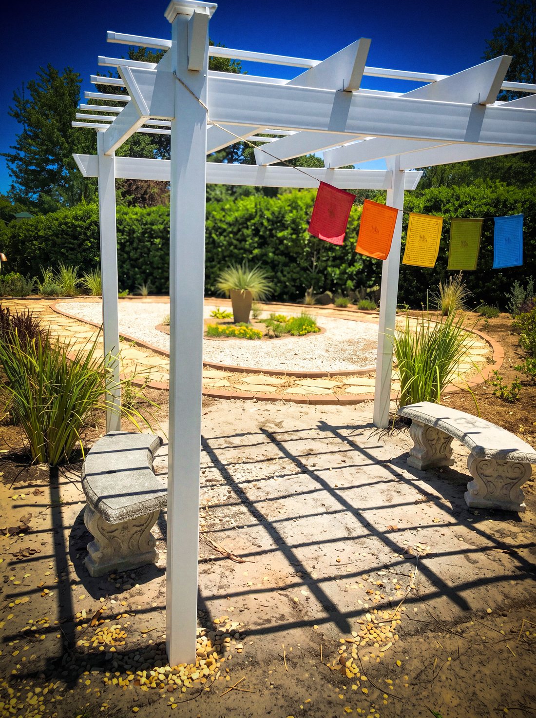The white pergola in the TriUU Memorial Garden, with colourful prayer flags and two stone benches on the gravel beneath it.
